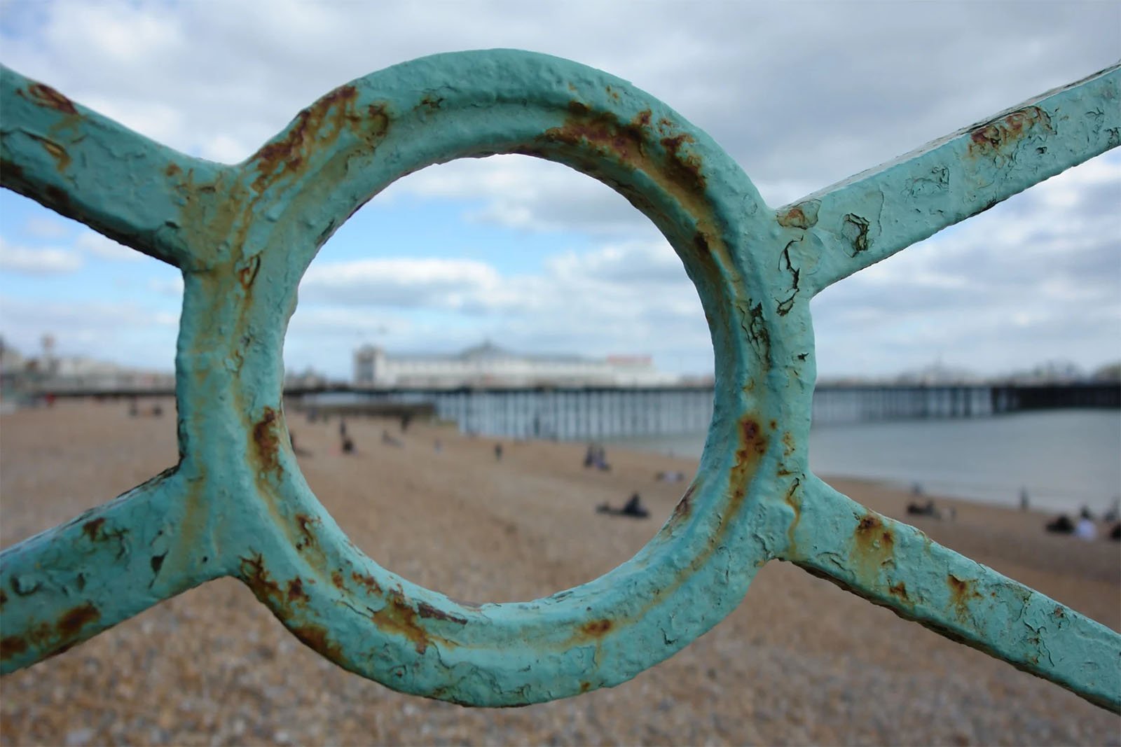 Close-up view of a weathered turquoise railing with a circular pattern framing a distant pier, sandy beach, and scattered visitors under a cloudy sky.
