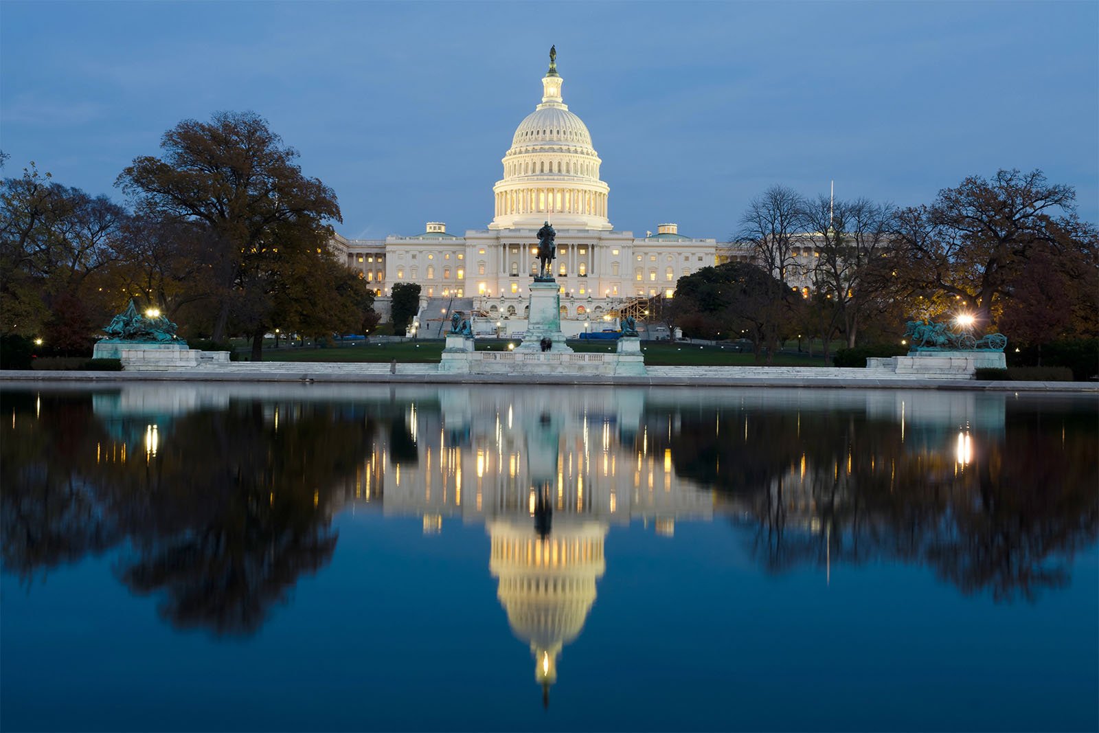 The united states capitol building illuminated at night, reflected perfectly in the calm waters of a pool, with surrounding trees in early twilight.