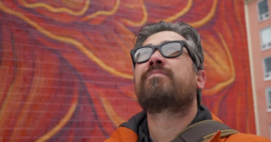 A man with a beard and glasses looking upwards, with a colorful, abstract mural in the background. he wears a brown jacket and carries a backpack strap over one shoulder.