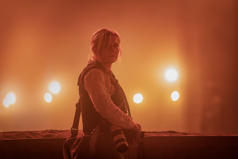 A woman with a camera around her neck stands in a misty, amber-lit scene, looking thoughtfully to the side. She carries a camera bag and wears a vest over a long-sleeve shirt.
