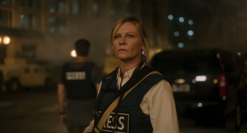 A woman wearing a press vest stands in a dimly lit urban area with emergency responders and vehicles in the background. She appears focused and serious.