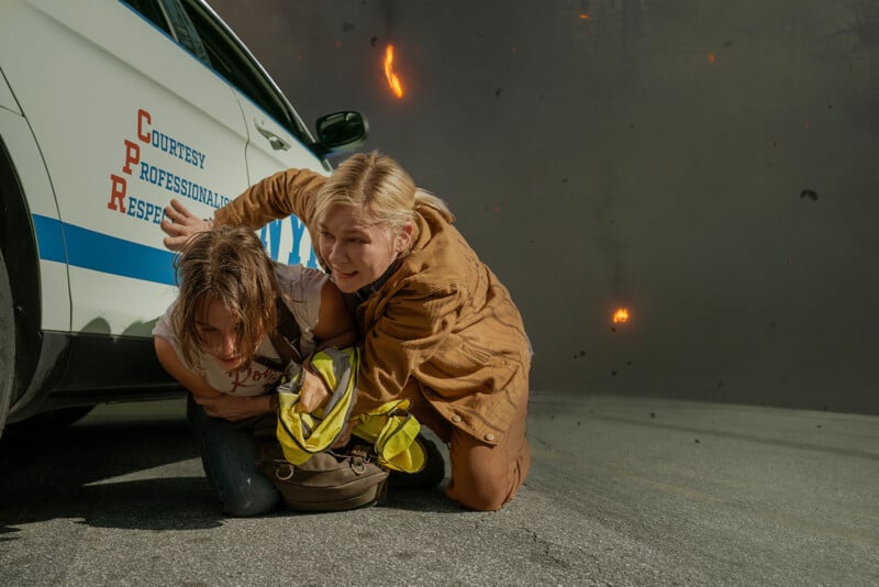 Two women crouch beside a white car with professional lettering, appearing distressed amidst a chaotic background with debris flying. One wears a brown jacket and the other a reflective work vest.