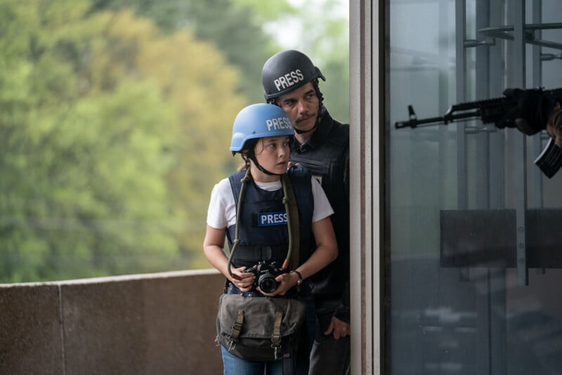 A male and a female journalist equipped with helmets labeled "press" and bulletproof vests stand near a building window, observing cautiously with a camera in hand.