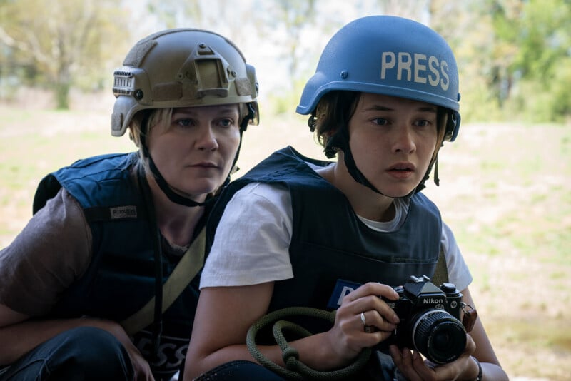 Two people wearing protective helmets and vests, labeled "press," attentively watch something in a park-like setting. one holds a camera, ready to photograph.
