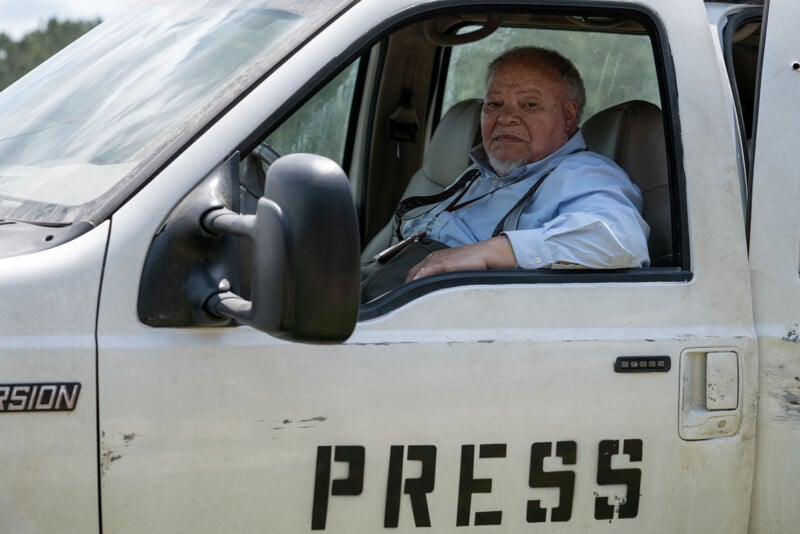 An elderly male journalist sits in a white van marked with "press" on the door, looking out the window with a serious expression. The van appears weathered and dusty.