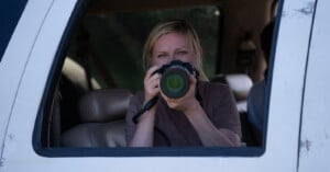 A woman with blonde hair sits in a vehicle's driver seat, looking through the viewfinder of a dslr camera aimed out the window. The vehicle's interior is visible.