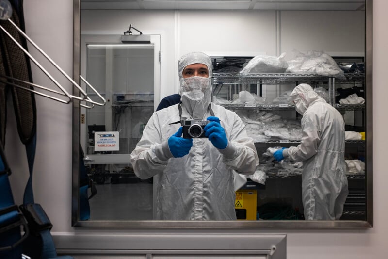 A man in a cleanroom suit holds a camera, seen reflected in a mirror, with shelves of similar suits and equipment in the background.