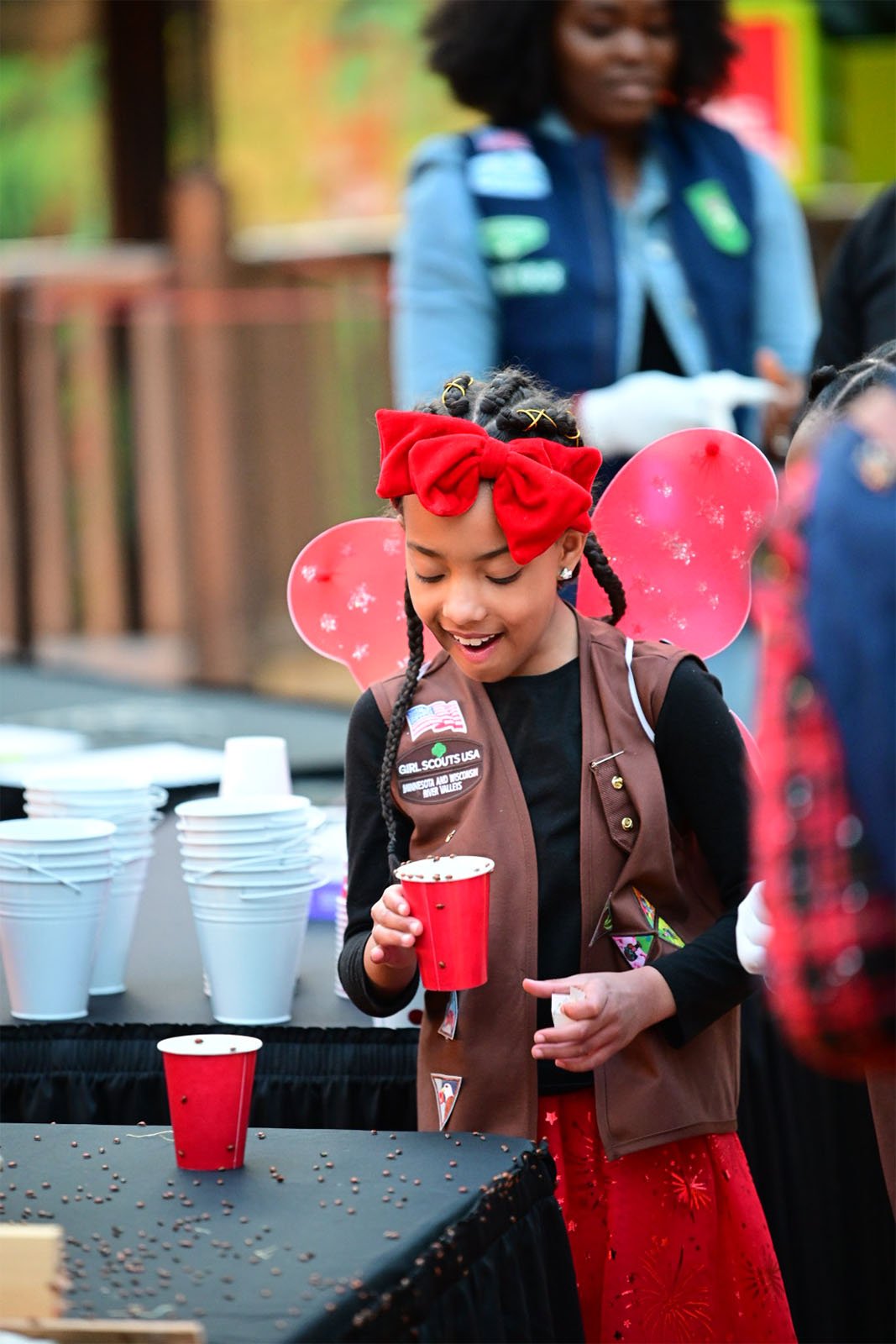 A young girl wearing a red headband and girl scouts vest smiles while holding a red cup at an outdoor event, with other people and a table with cups in the background.