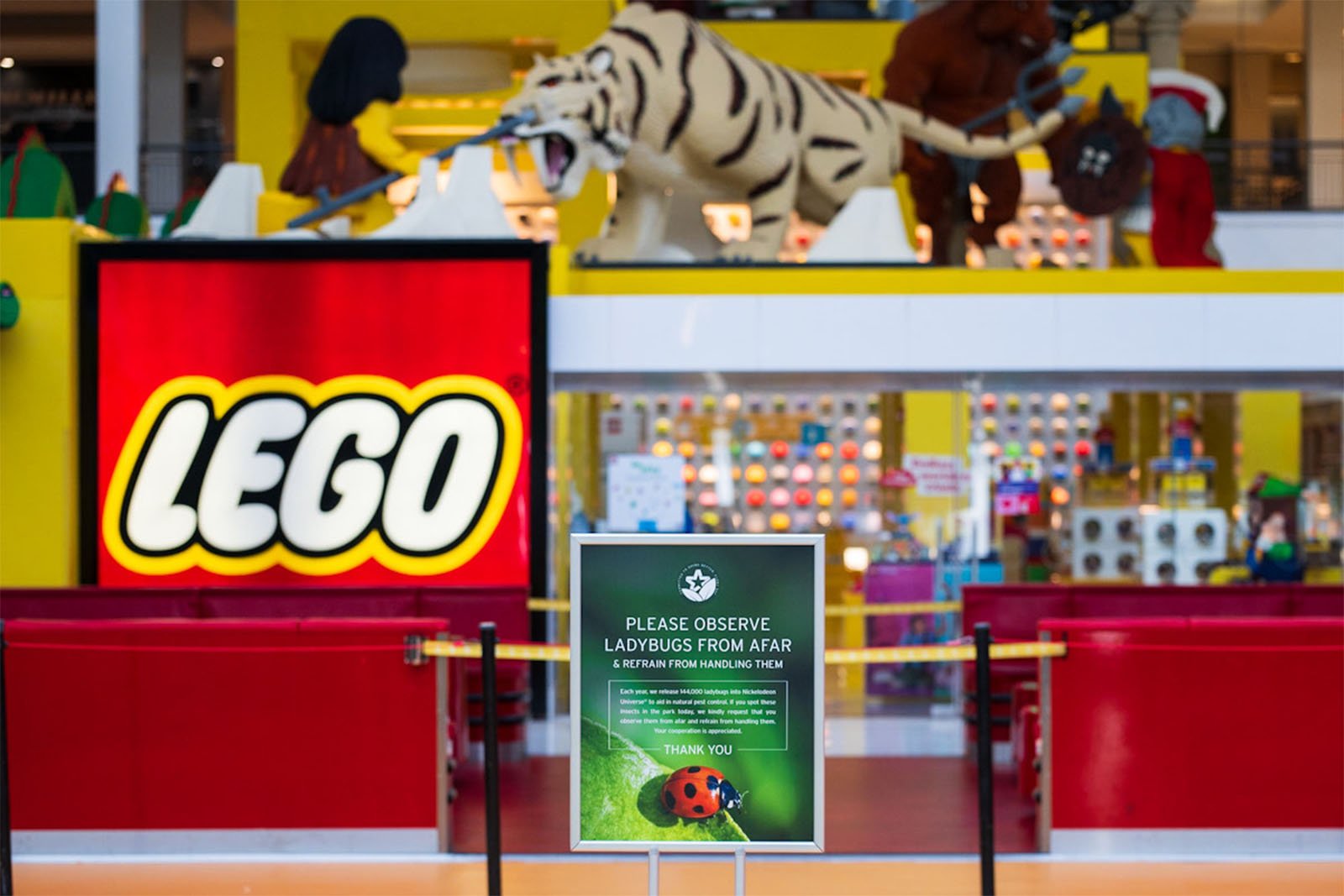 Bright lego store entrance displaying large lego logo with models of animals like a tiger and bear. foreground has a sign requesting to observe ladybugs from afar for a scavenger hunt.