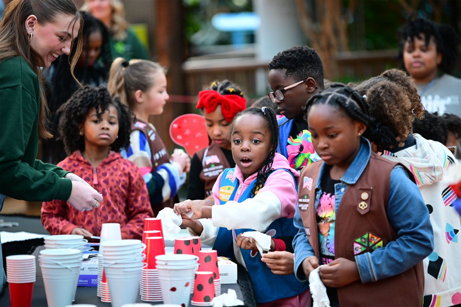 A group of excited children, some wearing scout uniforms, gather around a table with cups as a smiling woman assists them during an outdoor event.