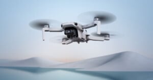 A white drone with a camera flies over a tranquil landscape with gentle rolling hills and a serene blue sky in the background.