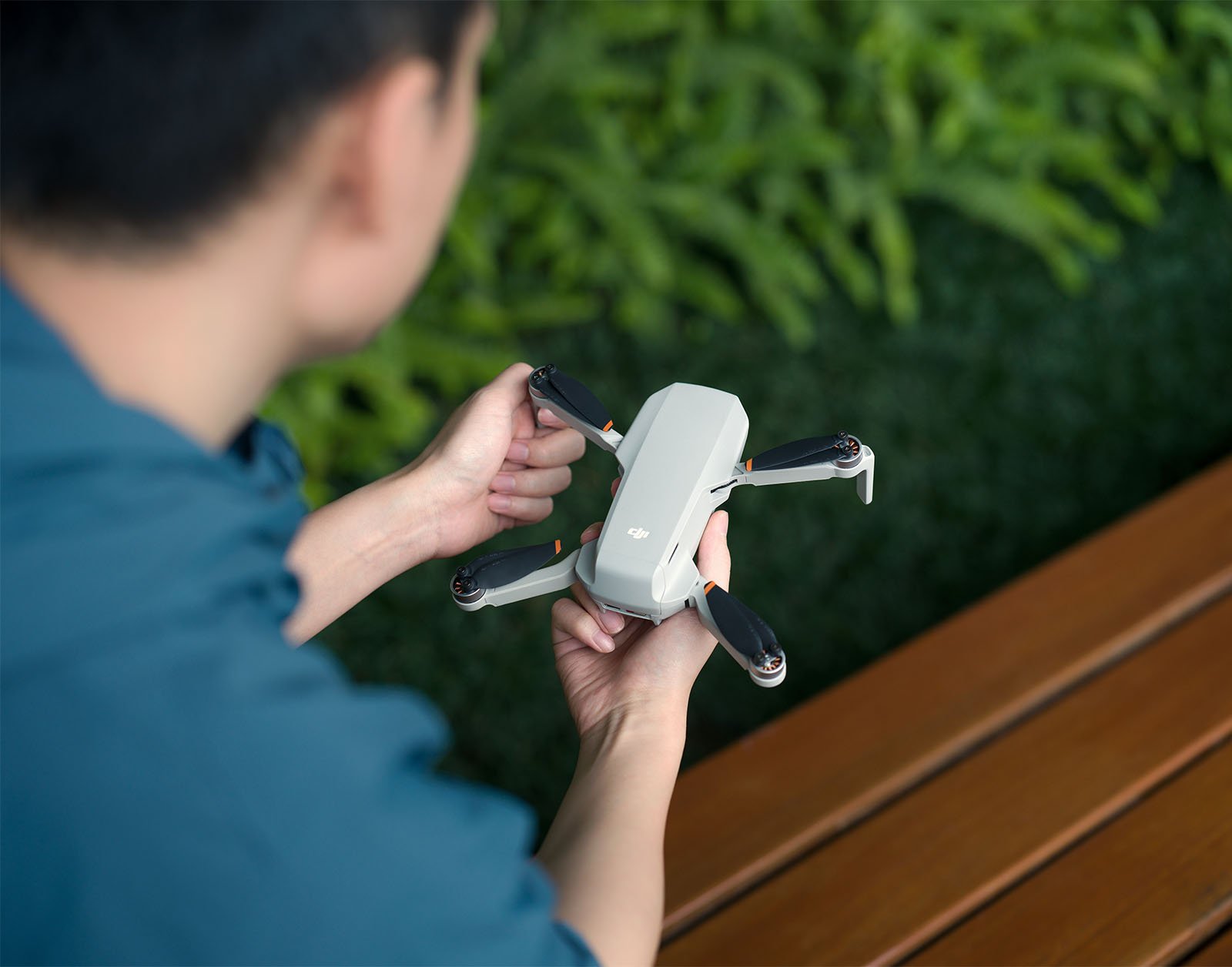A person holding a small white drone with extended propellers, preparing for launch, sitting on a bench with a lush green hedge in the background.