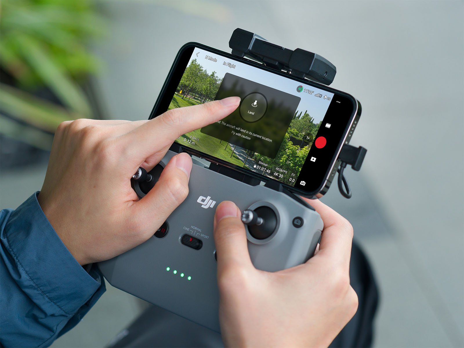 Hands of a person using a drone controller with a smartphone attached, displaying a camera view of greenery on the screen.