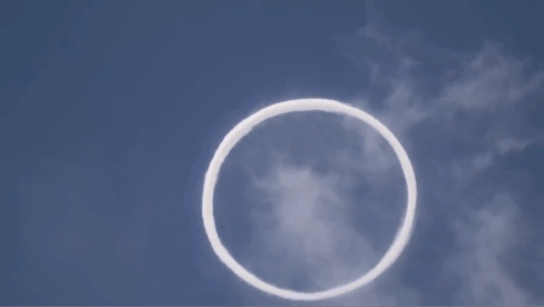 A white circular smoke ring floats against a clear blue sky, creating a striking visual contrast.