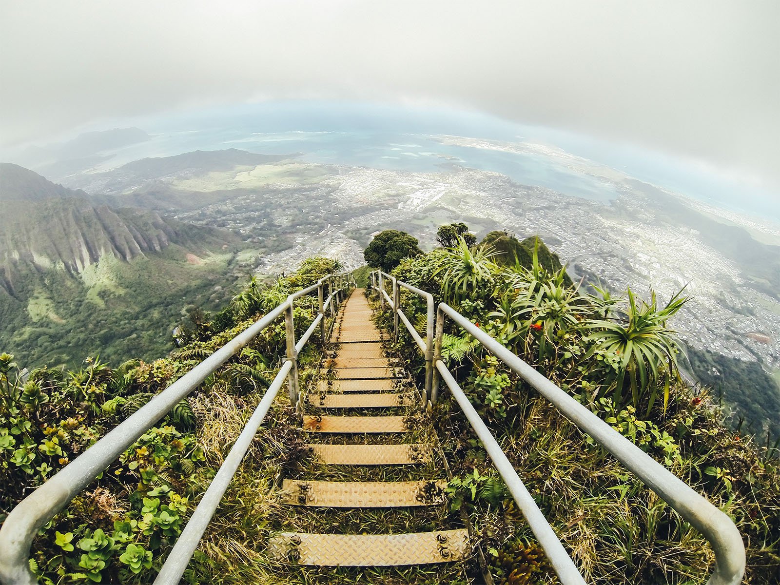 A wide-angle view from the top of a mountain staircase descending into a lush landscape with coastal views, urban areas, and overcast skies.