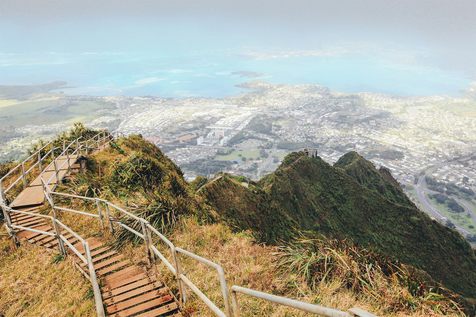 Aerial view from a mountainous hiking trail with a wooden staircase leading down towards a lush valley, overlooking a coastal town and bright blue sea in the distance.