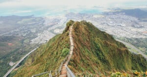 A narrow hiking trail with wooden steps along a steep, lush green mountain ridge, overlooking a panoramic view of a coastal city and ocean.