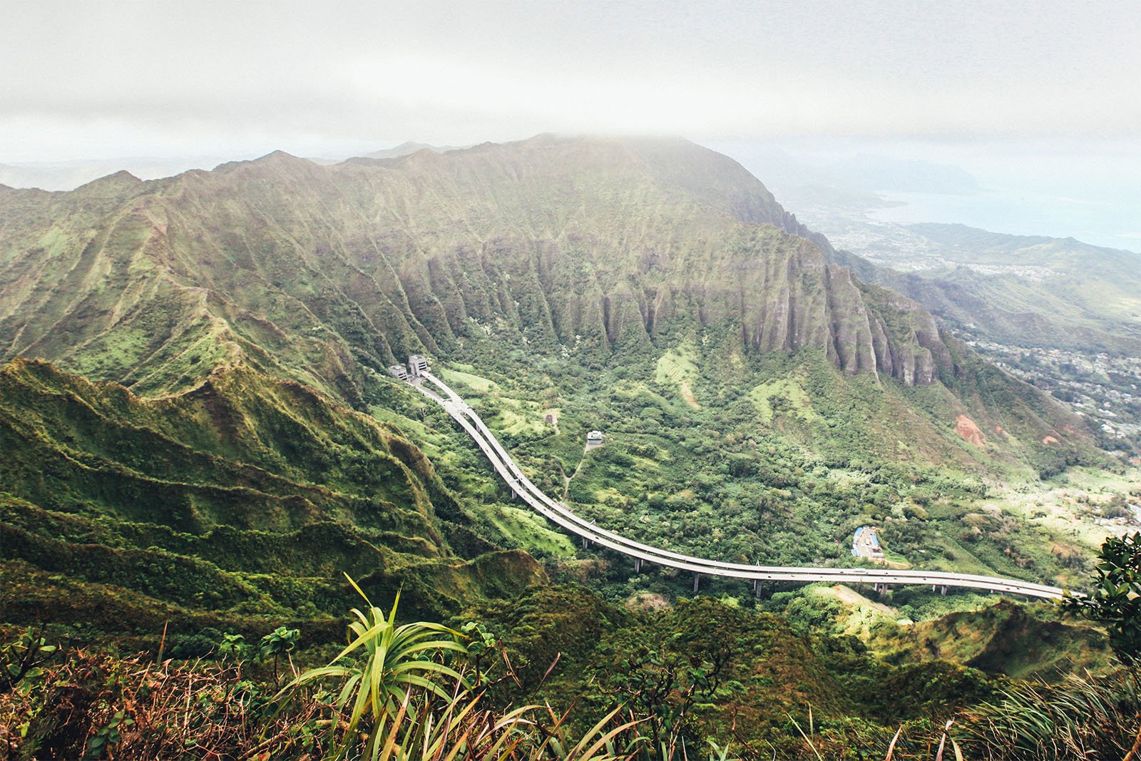 A winding road cuts through lush, green mountain terrain, overlooking distant ocean views under a cloudy sky.