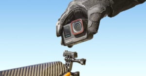 A gloved hand holding an Insta360 Ace Pro action camera, pointed toward a camera mount set against a clear blue sky.