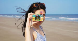A woman holds the LomoApparat Fluffy Omelet Edition at a beach.