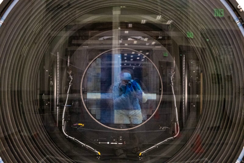 A person in a cleanroom suit taking a photograph through the lens of the LSST camera looking directly into the sensor, reflected off it. multiple reflections are visible in the glass.