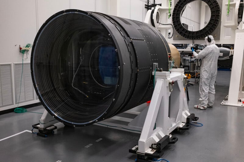 Two technicians in clean suits work on a large cylindrical space telescope component in a high-tech lab environment.