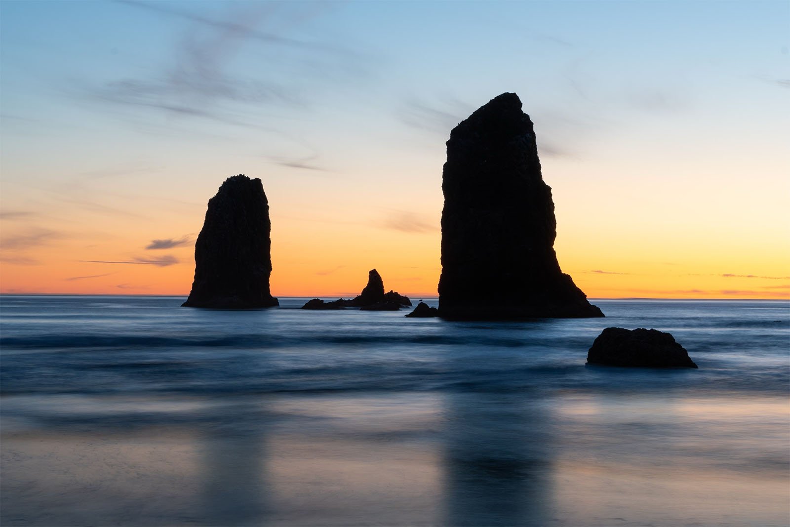 A serene sunset at the beach showing three large rock formations rising from the sea against a vivid orange and blue sky, with the ocean in the foreground appearing smooth and misty.