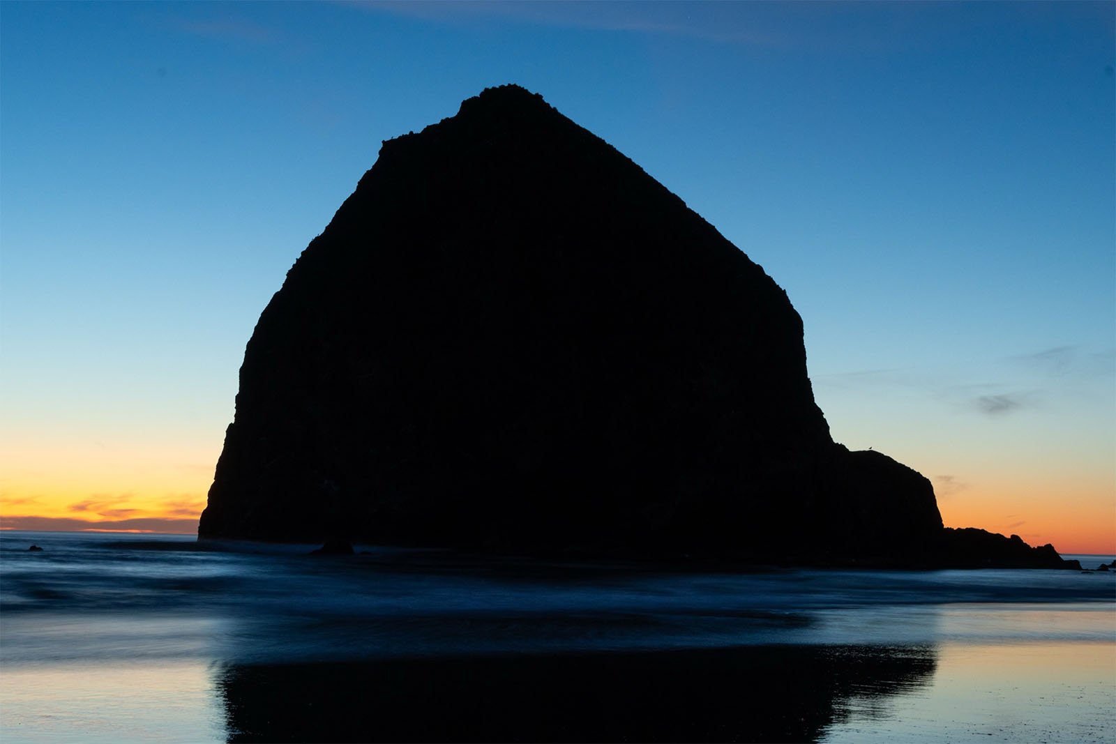 Silhouette of a large rock formation on a beach at sunset, with an orange and blue sky reflected on the wet sand.