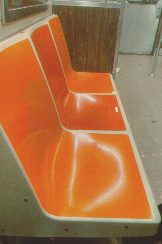 An orange subway seat made of plastic, with a shiny surface reflecting light, situated in a carriage with metal poles and wooden paneling visible in the background.