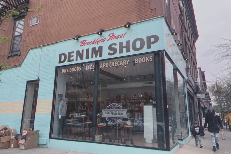 Corner view of a shop with the signage "Brooklyn's Finest Denim shop" featuring large windows, displaying various items inside, with pedestrians walking by.