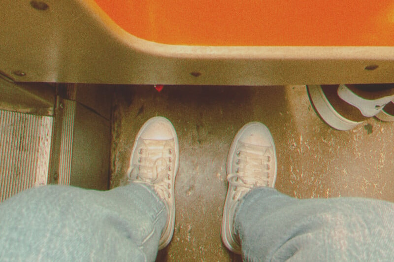 A person's feet in white sneakers and blue jeans viewed from above while sitting on public transportation, with an orange seat nearby and another person's foot visible.
