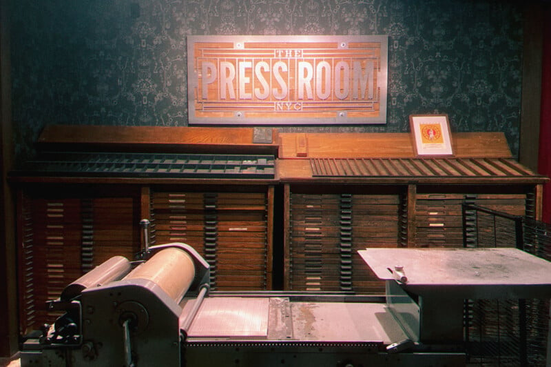 A vintage printing press setup featuring an old press machine, various type cases, and a lit sign reading "The Press Room NYC" on a patterned wall in a dimly lit room.