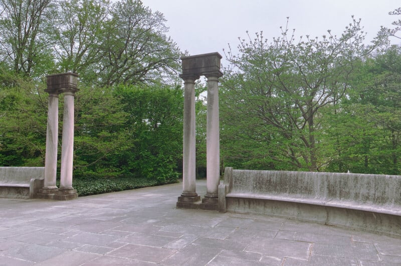 Two ornate stone columns flank an opening on a weathered stone terrace, surrounded by lush green trees in a serene park-like setting.