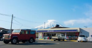A striking view of mount fuji in the background with a clear blue sky, visible behind a convenience store and two vintage vehicles parked outside.