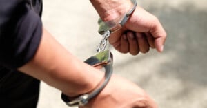 A close-up of a person's wrists handcuffed, with a focus on one hand and the metal cuffs. the person is wearing a dark blue shirt, and the background is a blurred light gray surface.