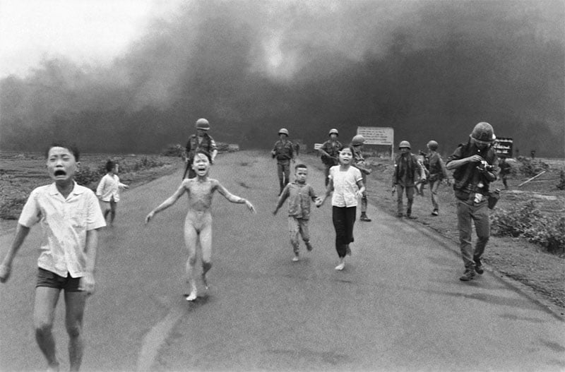 A historical black-and-white photo showing a group of children and adults running on a road. one child appears particularly distressed. smoke billows in the background, and soldiers walk behind.
