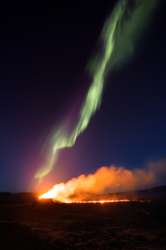 A stunning display of the northern lights in a dark sky above a vibrant, expansive ground fire emitting glowing flames and smoke.
