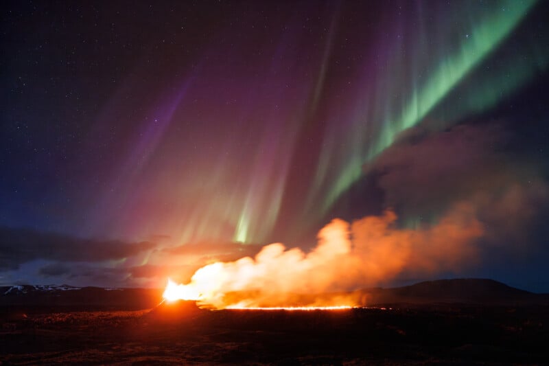 A dramatic scene where the northern lights illuminate the sky with green and purple hues above an erupting volcano spewing red lava and billowing smoke.