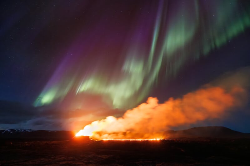 A vibrant display of the northern lights in hues of green and purple over a fiery volcanic eruption, casting an orange glow against a dark night sky.