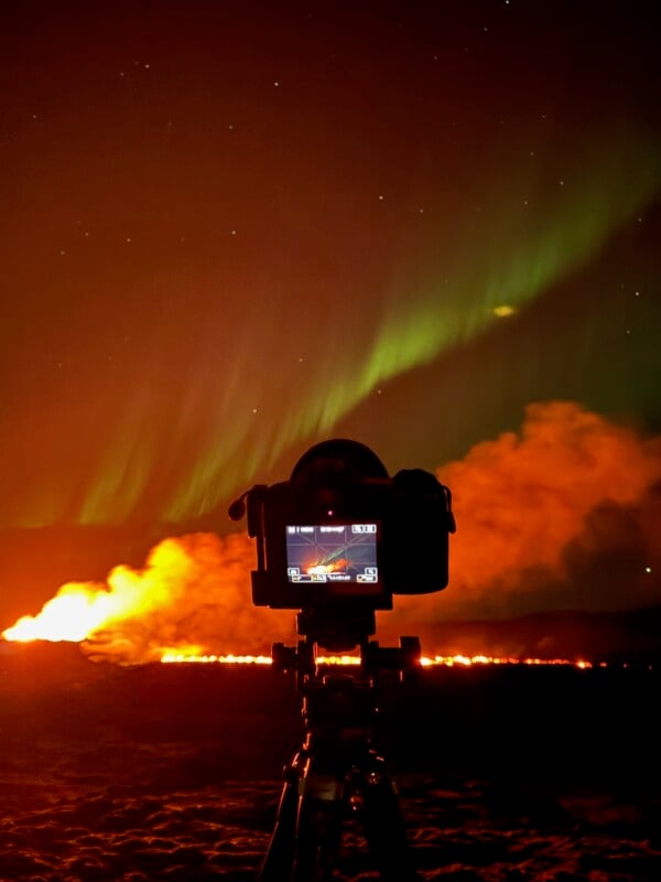 A camera on a tripod capturing the aurora borealis and a volcanic eruption at night, with a fiery lava flow and a green-lit sky visible on its display screen.