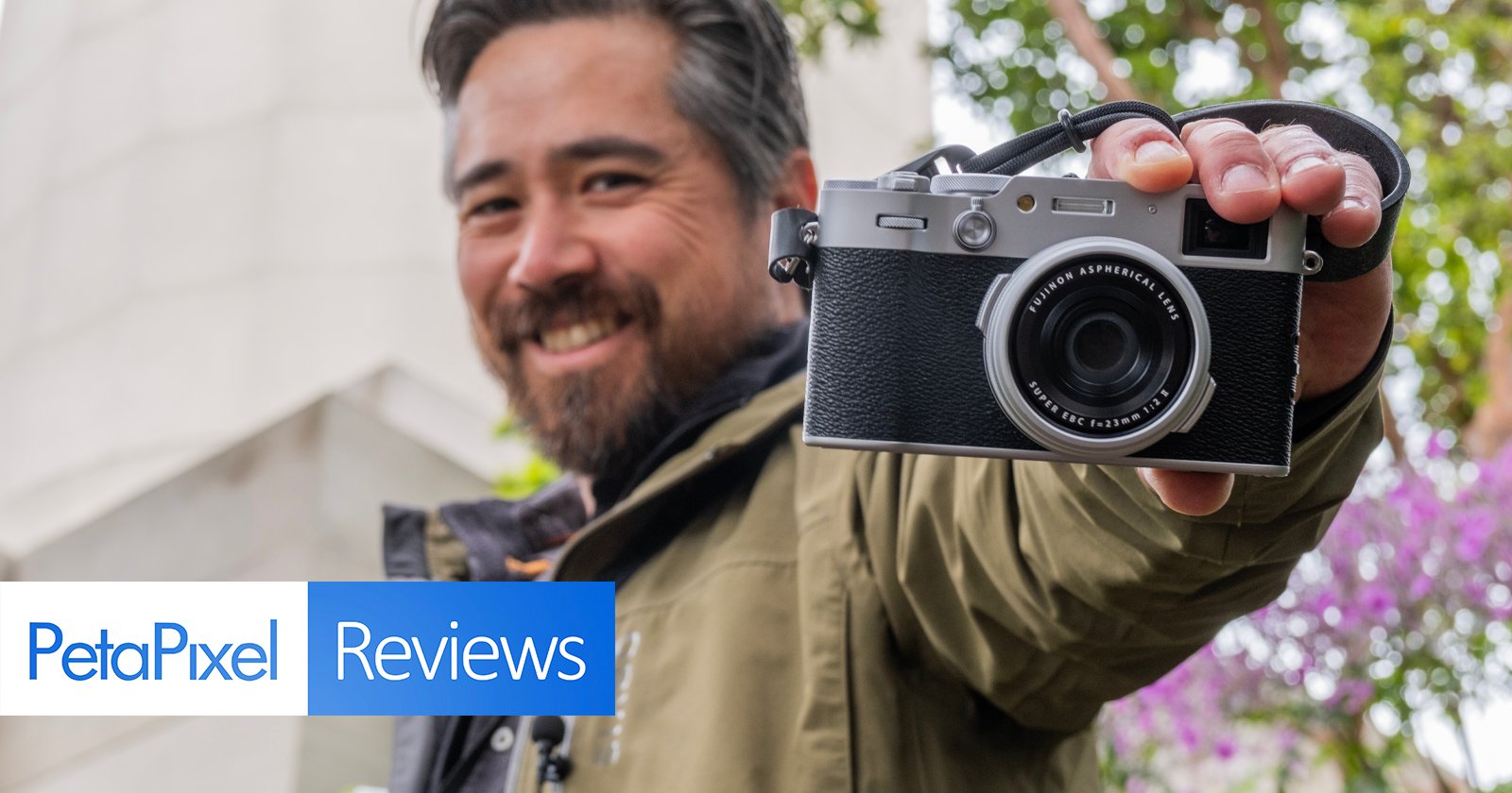 A smiling man holding a camera in front of him, focused on the lens, with "petapixel reviews" overlay text, in a garden setting.