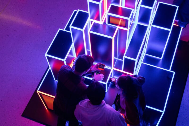 Top view of three individuals interacting with a large, glowing neon geometric installation, using a Rabbit R1. Bright pink and blue lights illuminate the area.
