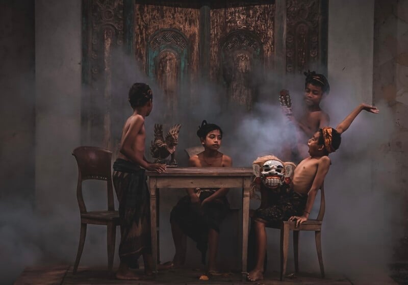 Four children engage in a mystical, theatrical scene in a dimly lit room with rustic decor, featuring masks and expressive gestures around a wooden table.
