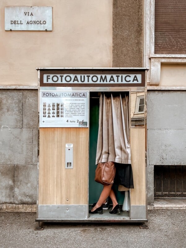 A woman stands partially inside a small photo booth labeled "fotoautomatica" on a city street, with only her legs visible from the knee down.