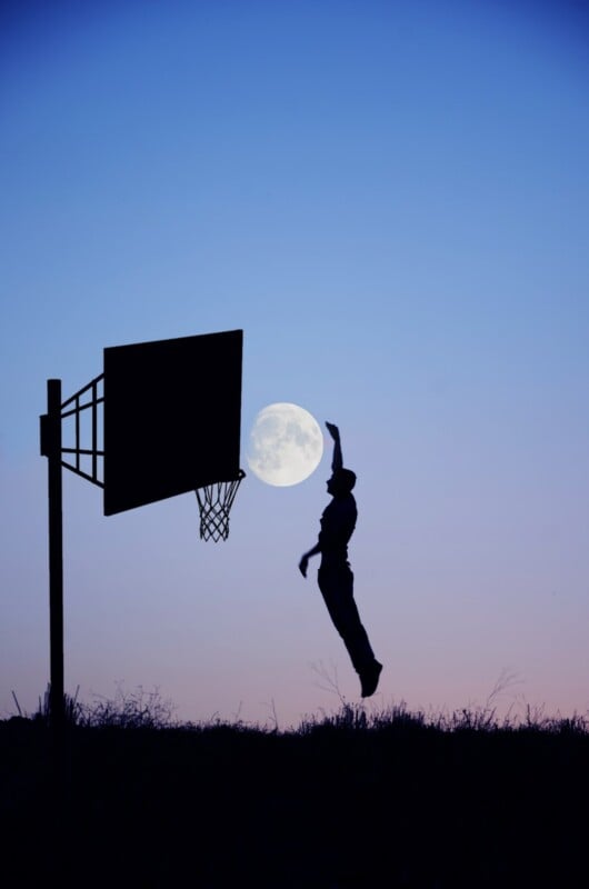 A silhouette of a person shooting a basketball at an outdoor hoop against a twilight sky, with a full moon perfectly aligned behind the hoop.