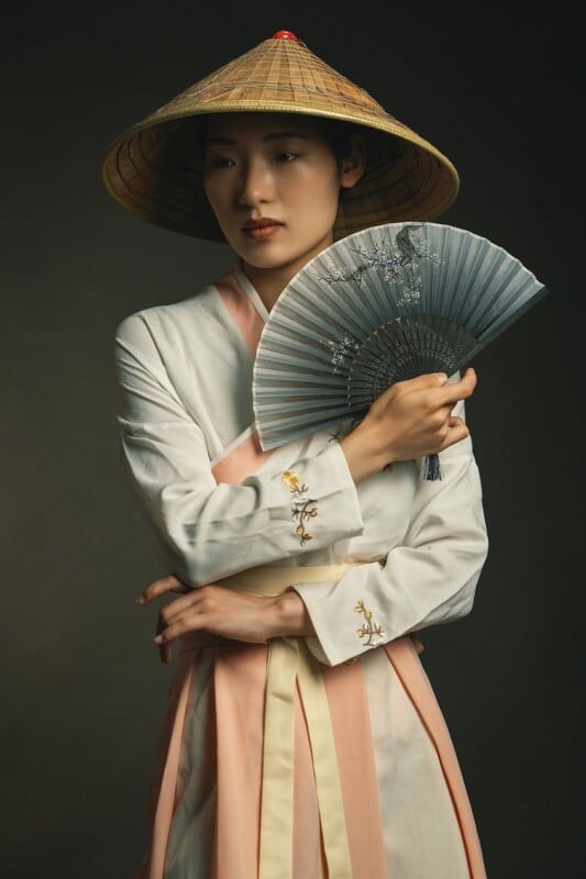 A woman dressed in a traditional east asian hanbok and conical hat holds an intricate silver fan, looking contemplative against a dark background.
