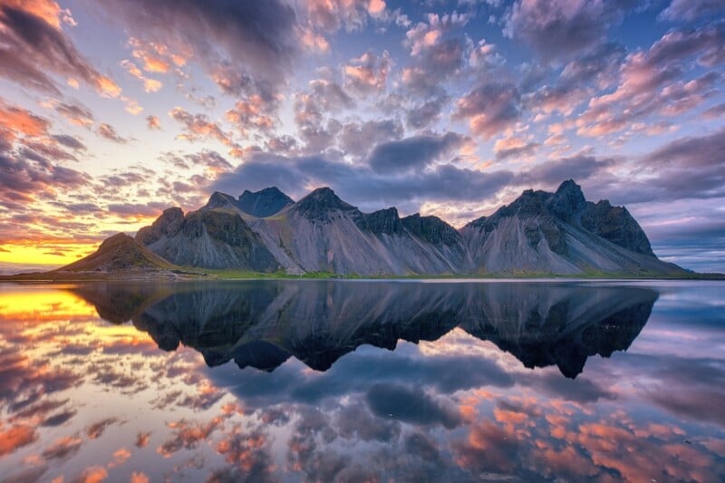 A breathtaking view of a mountain range reflected in a tranquil lake at sunrise, with colorful clouds scattered across a vibrant sky.