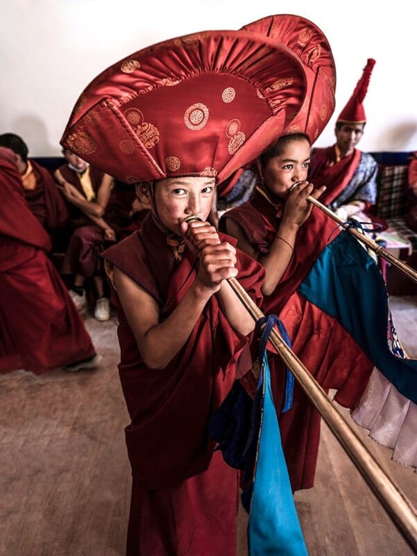 Two young monks in traditional attire play musical instruments in a room, with others seated in the background. the focus is on the monk in the foreground wearing a large red ceremonial hat.