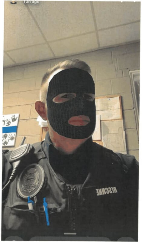 A police officer wearing a black balaclava, sitting in an office environment with visible name patches and badge on the uniform.