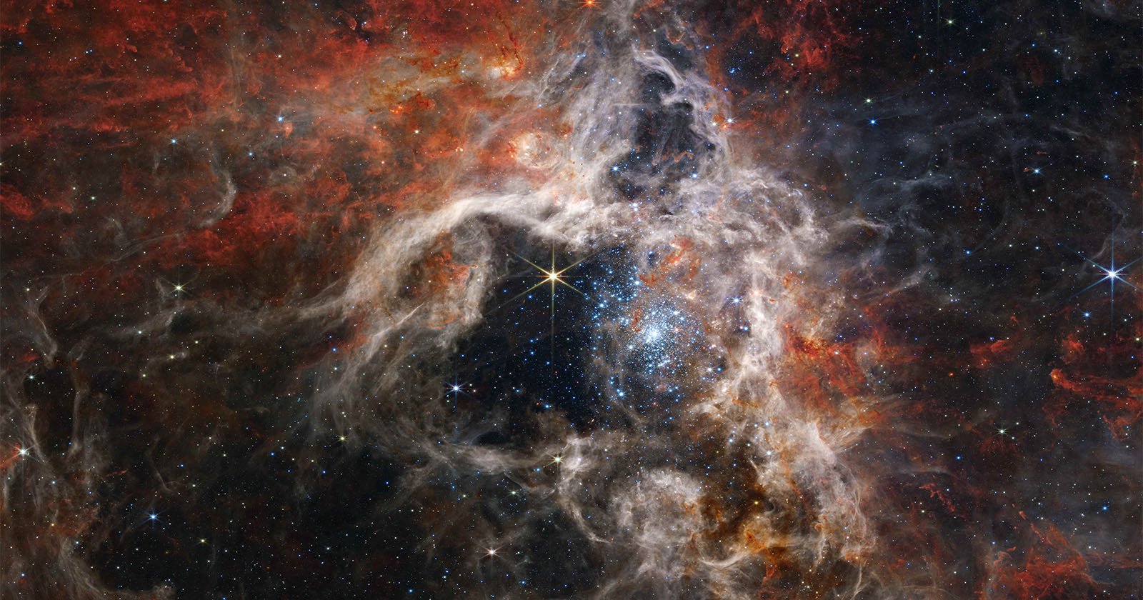 A vibrant cosmic scene featuring a cluster of bright stars surrounded by intricate networks of reddish-orange gas and dust nebulae, set against a dark space background.